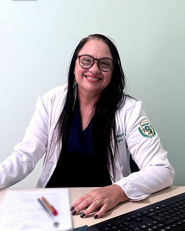Enfermeira Luciane Sampaio sorrindo, de jaleco branco, sentada à mesa de atendimento